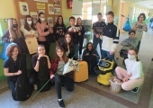 Szesnaście osób z klasy 7a na tle gazetki Samorządu Uczniowskiego z żółtą walizką i żółtym odkurzaczem w centrum zdjęcia. Dwie dziewczyny trzymają reklamówki na kijkach.